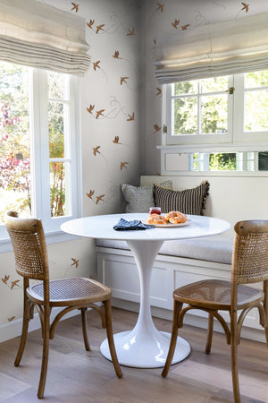 The Sparrow brown wallpaper by Rylee and Cru is in a dining nook with a white sculptural dining table, brown woven chairs that are surrounded by large colonial style windows.