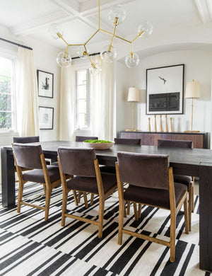The Bobbi gold branching chandelier with glass orb bulbs is mounted in a dining room with brown velvet dining chairs, a black wooden dining table, and a black and white striped rug