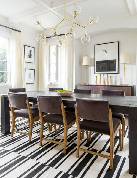 #color::gold | The Bobbi gold branching chandelier with glass orb bulbs is mounted in a dining room with brown velvet dining chairs, a black wooden dining table, and a black and white striped rug