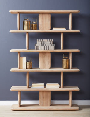 The Nera natural solid wood sculptural bookcase sits against a blue wall with books and brass containers stacked within its shelving.