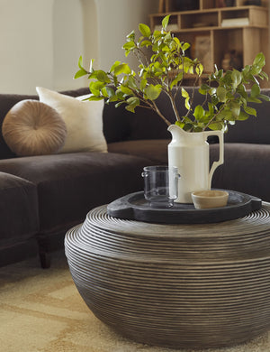The Lylah round wooden black tray sits atop a round wooden coffee table in a living room with a brown velvet sofa and a white vase