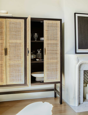The Hannah black mango wood cabinet with cane doors sits in a living room with one door open holding a white centerpiece bowl and hosting serveware.