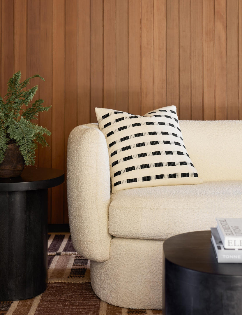 #color::onyx | Bertu onyx black pillow with a woven dash pattern by Bolé Road Textiles sits on a cream boucle rounded sofa in a room with wooden paneled walls