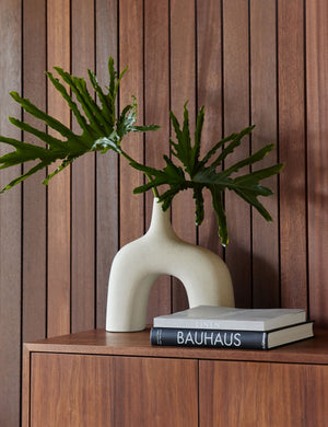 The Leonor larger sculptural arched matte white ceramic Vase sitting atop a wooden sideboard with a stack of books in front of a wood paneled wall