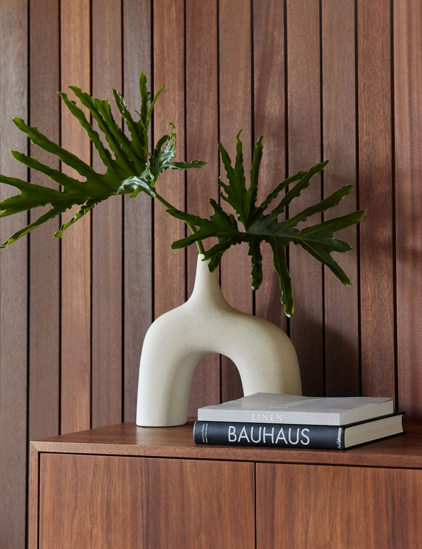 #color::white #size::large | The Leonor larger sculptural arched matte white ceramic Vase sitting atop a wooden sideboard with a stack of books in front of a wood paneled wall