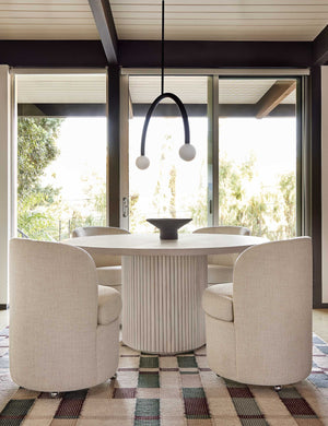 The Rutherford white-washed acacia wood round dining table with pedestal base sits in a dining room surrounded by four linen dining chairs atop a beige patterned carpet with red and green accents.