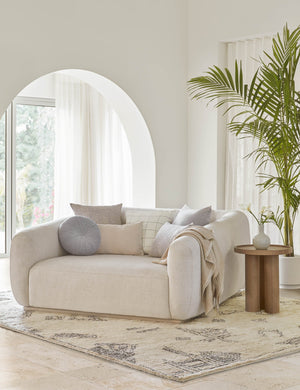 The Monroe ice blue velvet round pillow sits on a white lounge chair in a living room with a plush patterned rug