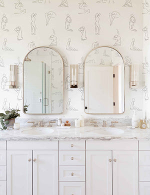The Minimalist white and black nude wallpaper is in a bathroom with two silver framed arched mirrors and double sinks with marble counter tops