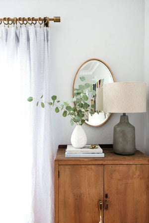 The Merci gold framed oval mirror is hung against a white wall behind a wooden side table and a white textured vase