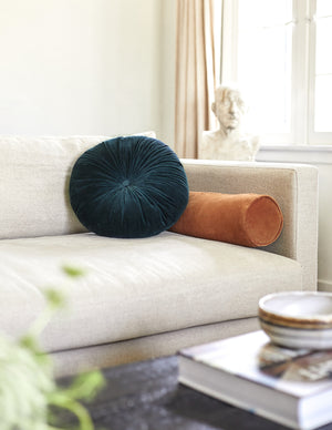 The Sabine burnt orange velvet cylindrical bolster pillow sits on a gray linen sofa in a living room with a sculptural object and a deep blue velvet disc pillow