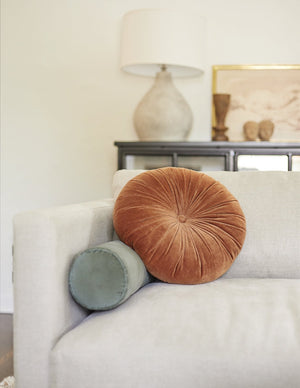 The Monroe burnt orange velvet round pillow sits on a white sofa with a green velvet lumbar pillow