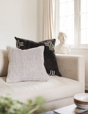 The Nico black handmade mudcloth throw pillow with an alternating x-motif pattern sits on a natural toned sofa with a black and white patterned throw pillow in a living room