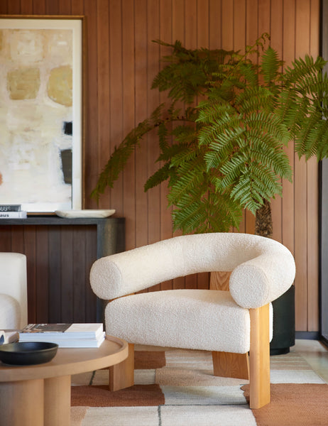 #color::cream | The Celeste honey wood accent chair with wishbone frame sits in a retro living room with wood paneled walls, a black planter, and a round wooden coffee table.