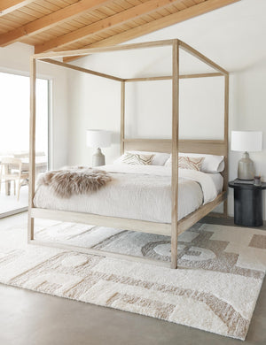 The Oasis plush geometric neutral-toned rug by Élan Byrd lays in a bedroom with a white wooden canopy bed, a gray sheepskin throw, and black wooden nightstands