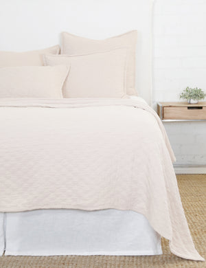 The Ojai light pink Cotton Matelassé Sham with a diamond woven pattern by Pom Pom at Home lays on a white linen framed bed in a bedroom with a white brick wall, a jute rug, and a wooden nightstand