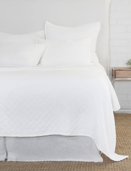 #color::white #size::king #size::queen #size::twin | The Ojaj white cotton matelassé coverlet by pom pom at home lays atop a white linen framed bed in a bedroom with a jute area rug, a white brick wall, and a wooden nightstand