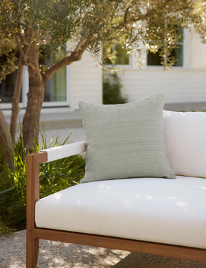 Moss green Milan indoor and outdoor square pillow with a linear pattern by Sunbrella sits on a white cushioned sofa in an outdoor space