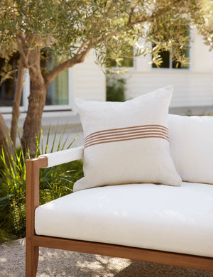 Katya Indoor and Outdoor square cream Pillow with rust brown stripes in the center sits on a white cushioned sofa in an outdoor space