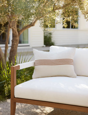 Katya Indoor and Outdoor lumbar cream Pillow with rust brown stripes in the center sits on a white cushioned sofa in an outdoor space