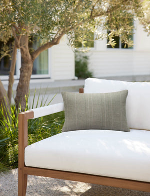 Moss green Milan indoor and outdoor lumbar pillow with a linear pattern by Sunbrella sits on a white cushioned sofa in an outdoor space