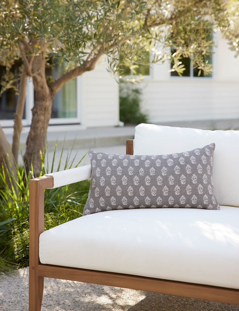 | The Montrose Indoor and Outdoor gray, paisley patterned Lumbar Pillow by Sunbrella for Lulu and Georgia lays on a white cushioned sofa in an outdoor space