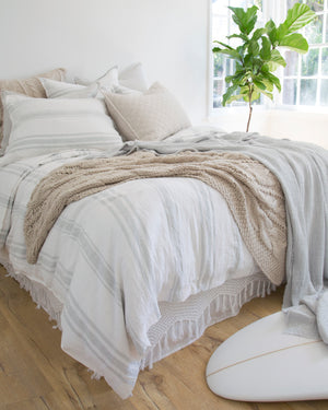 The Jackson Linen white and ocean striped Duvet by Pom Pom at Home lays on a bed in a bedroom with textured throw pillows, bright windows, and hardwood floors