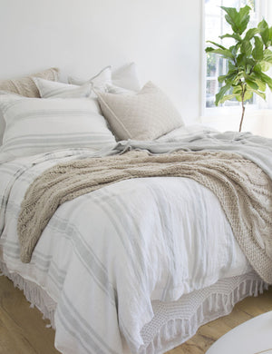 The Jackson Linen white and ocean striped Sham by Pom Pom at Home lays on a bed in a bedroom with textured throw pillows, bright windows, and hardwood floors