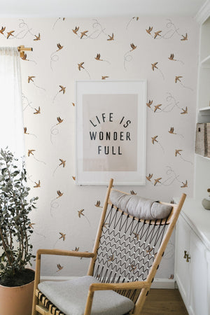 The Sparrow brown wallpaper by Rylee and Cru is in a room with a graphic wall art mounted against it behind a natural vase and a woven rocking chair with gray cushioning.