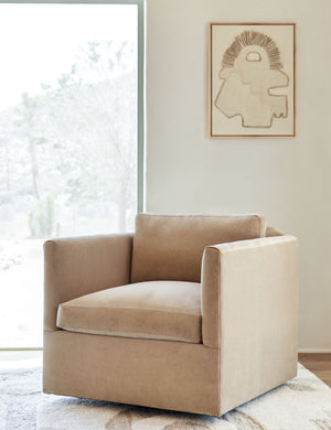 Lotte camel velvet swivel chair sits in a room with floor to ceiling windows, and an neutral-toned abstract wall art