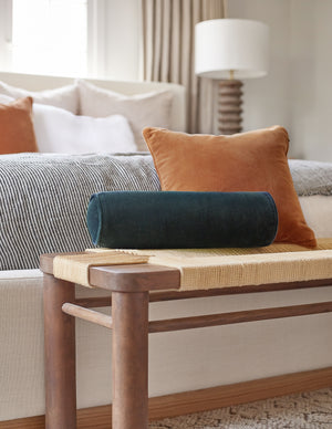 The Sabine navy velvet cylindrical bolster pillow sits atop a woven bench with a burnt orange velvet throw pillow in a bedroom at the end of a natural linen framed bed