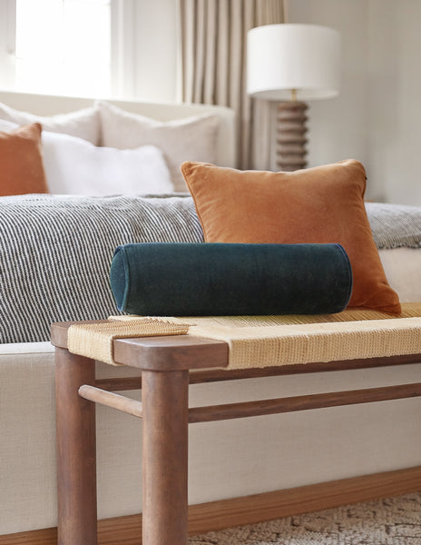 #color::navy | The Sabine navy velvet cylindrical bolster pillow sits atop a woven bench with a burnt orange velvet throw pillow in a bedroom at the end of a natural linen framed bed