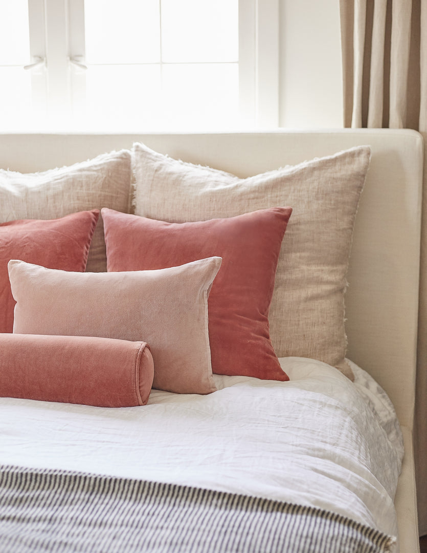 #color::coral | The Sabine coral velvet cylindrical bolster pillow sits on a white linen framed bed in a bedroom with striped linens and pink velvet throw pillows