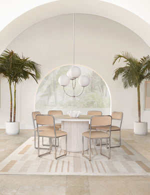The Rutherford white-washed acacia wood round dining table with pedestal base sits in an airy dining room surrounded by six light pink dining chairs atop a natural patterned rug with two palm trees and a large archway behind it.