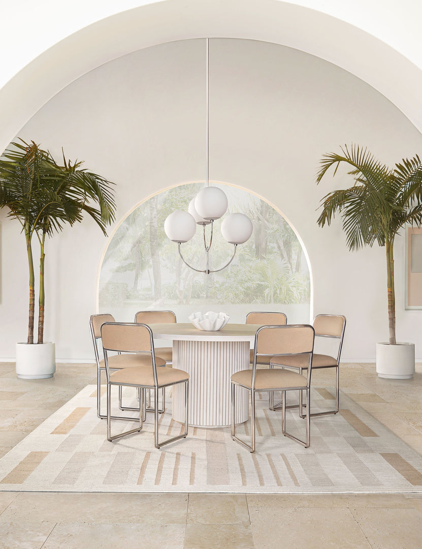 | The Rutherford white-washed acacia wood round dining table with pedestal base sits in an airy dining room surrounded by six light pink dining chairs atop a natural patterned rug with two palm trees and a large archway behind it.