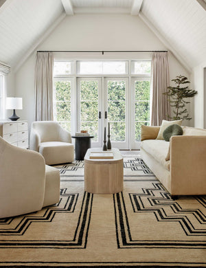 The Senna neutral hand-knotted wool area rug with black geometric pattern sits in a neutral living room with cream sofa, ivory accent chairs, and a light wood oval coffee table.