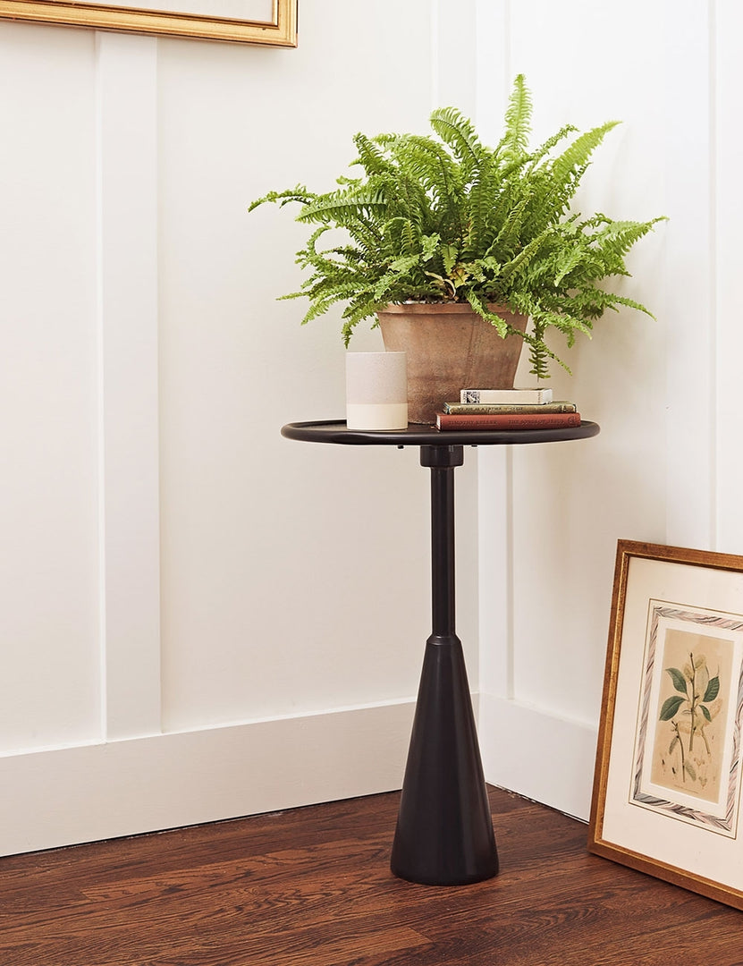 | The Arashi black metal side table with rounded top and sculptural base sits in the corner of the room with white accented walls, a terracotta planter, and a floral wall art