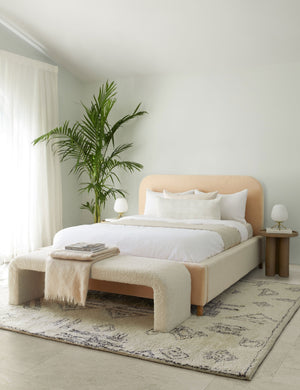 The Lucian white and rust lumbar pillow sits on a buff pink velvet framed bed with white linens that is atop a plush rug