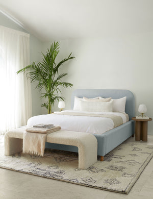 The Solene dove light blue velvet platform bed sits atop a plush patterned rug with a boucle cream bench and white linens