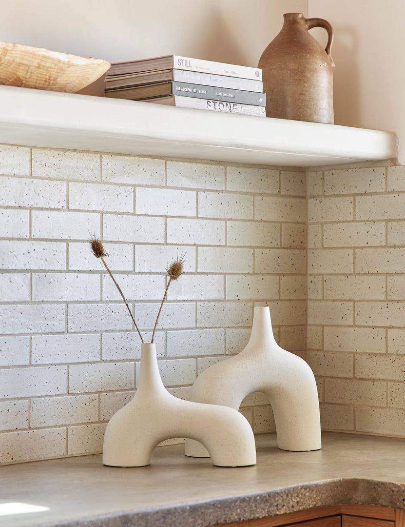#color::white #size::large | The Leonor sculptural arched matte white ceramic Vase in its small and large size sitting atop a granite countertop in a kitchen with a subway tile backsplash, a stack of books, and a wooden centerpiece bowl