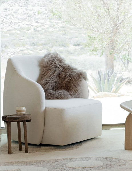 #color::latte | The Vale icelandic light gray sheepskin is laid in a living room atop a white accent chair with a wooden stool and coffee table