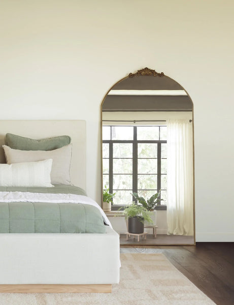 #color::gold | The Tulca gold curved standing mirror with flat bottom edge and traditional scroll detailing sits in a bedroom leaning against a white wall to the right up a natural linen upholstered bed with sage green bedding.