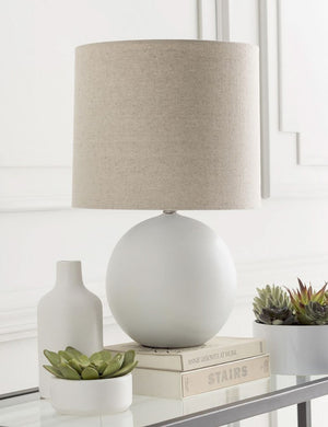 The Vivienne white ceramic table lamp with spherical base sits atop a glass sideboard with a white vase, cacti house plants, and a stack of books