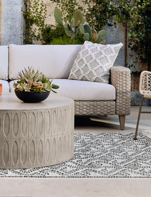 The Mal concrete indoor and outdoor round coffee table sits in an outdoor space with a gray and black patterned rug, a woven sofa with gray cushioning, and textured throw pillow
