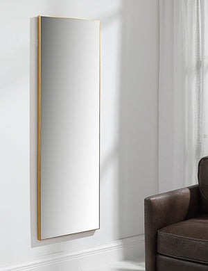 The Shea rectangular gold framed full length floor mirror hangs on a wall in a living room with a brown leather chair
