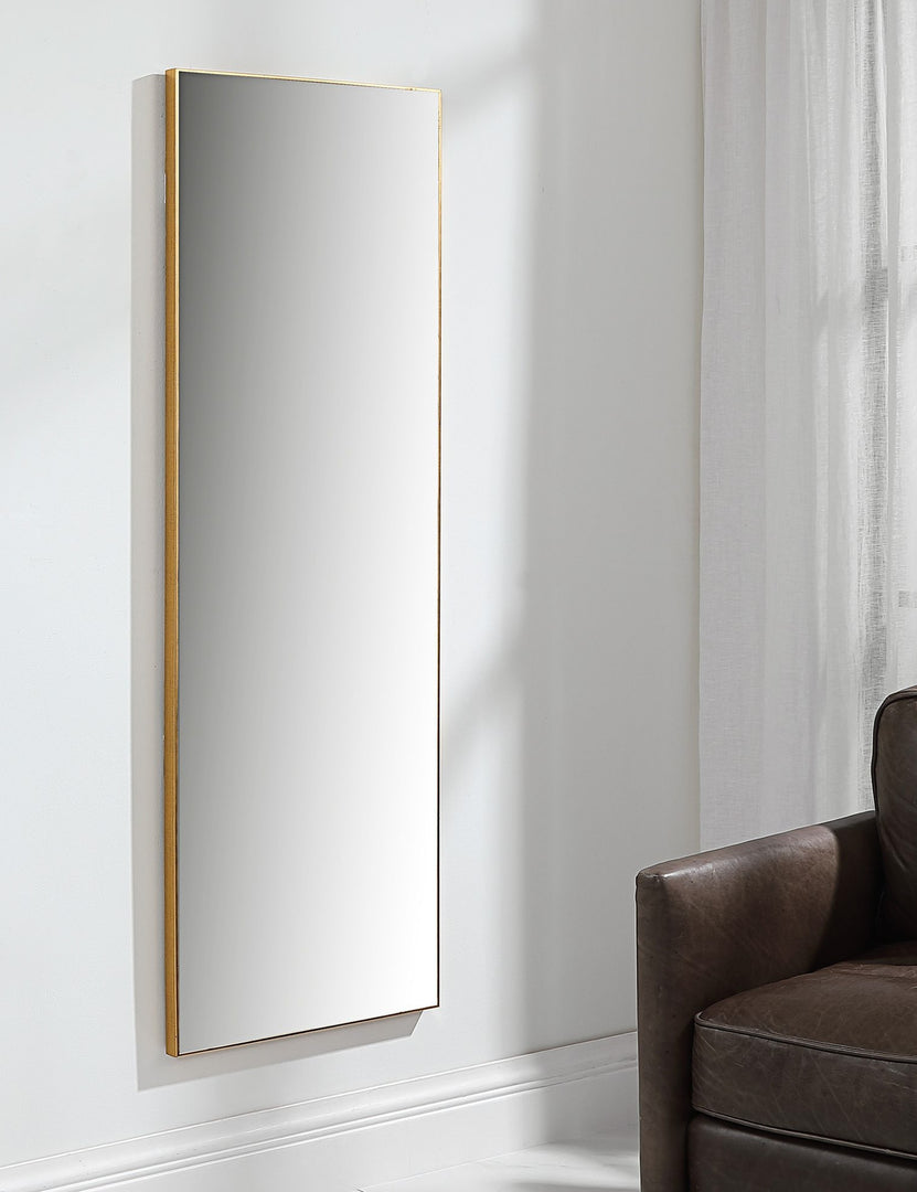 #color::gold | The Shea rectangular gold framed full length floor mirror hangs on a wall in a living room with a brown leather chair