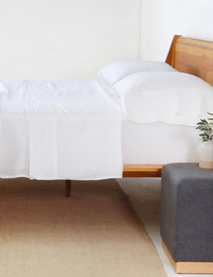 The white Linen Sheet Set by Pom Pom at Home lays on a wooden framed bed in a bedroom with a gray nightstand and a jute rug