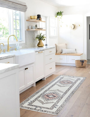 The Zehra hand-knotted ivory, gray and purple medallion wool-blend area rug sits in front of white kitchen cabinets with a light marble countertop and gold hardware.